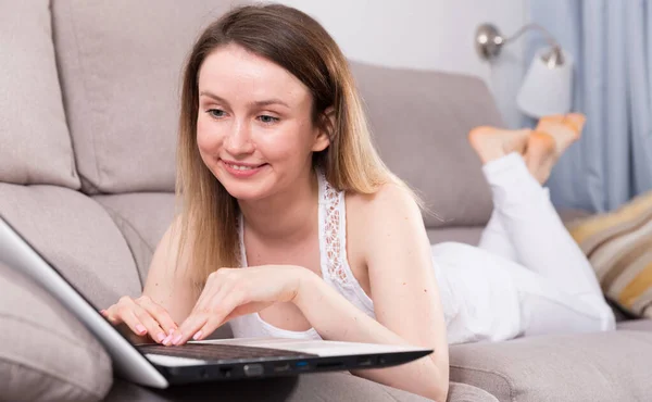 Mulher sorridente está trabalhando com laptop deitado no sofá — Fotografia de Stock