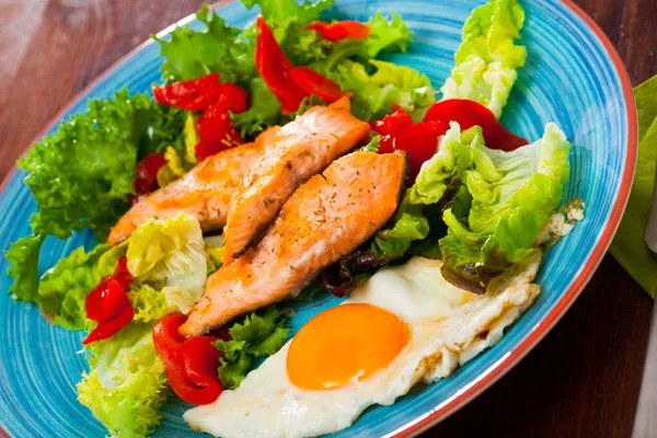 Изображение тарелки с жареной форелью, яйцом, овощами и зеленым салатом — стоковое фото