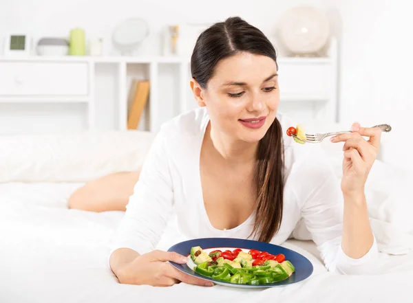 Pretty woman in white dress eating vegetable salad in bed — Stock Photo, Image