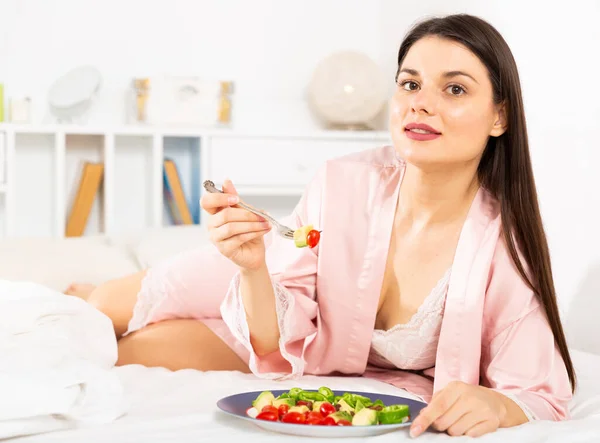 Woman in bathrobe holding fork and eating vegetable salad in bed — Stock Photo, Image