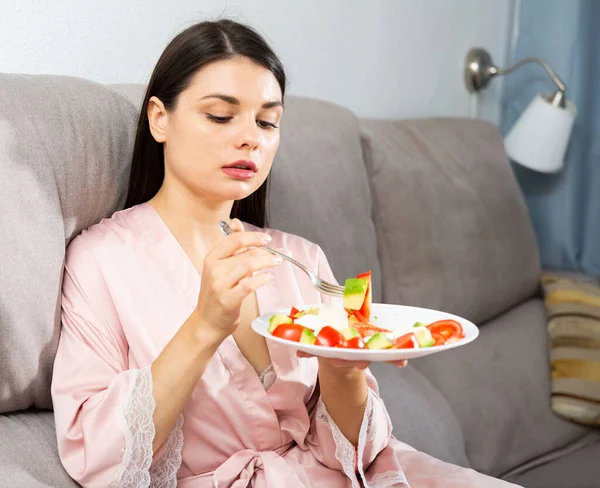 Zadowolona kobieta siedzi na kanapie w domu i je zdrowe jedzenie — Zdjęcie stockowe