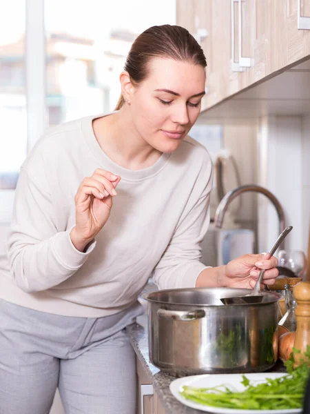 Iloinen naaras seisoo lähellä pöytää potin kanssa keittoa ja suolaa varten keittiössä — kuvapankkivalokuva