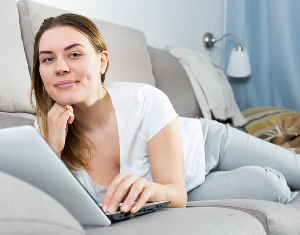 Chica alegre está acostado en el sofá y juguetón posando con el ordenador portátil —  Fotos de Stock