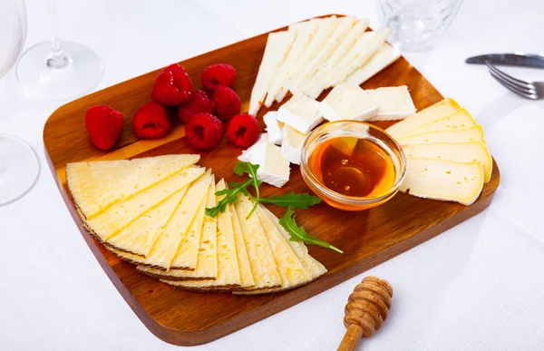 Slices of artisanal cheeses on wooden board with raspberries and honey — Stock Photo, Image