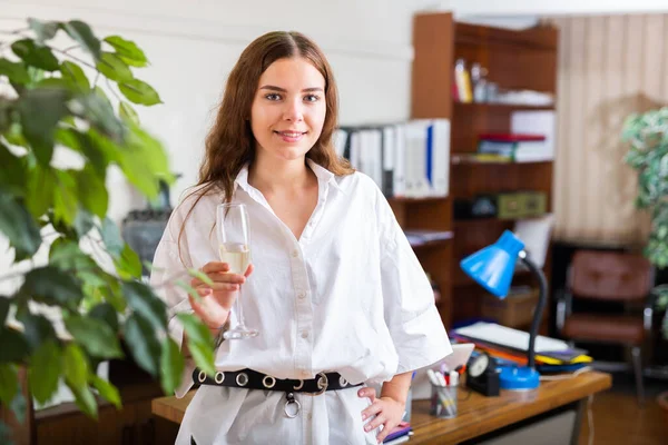Wesoła młoda kobieta ze szklanką szampana pozująca w biurze — Zdjęcie stockowe