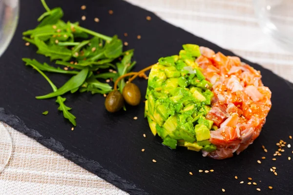 Salmon tartare with avocado served with capers and arugula on slate board — Stock Photo, Image