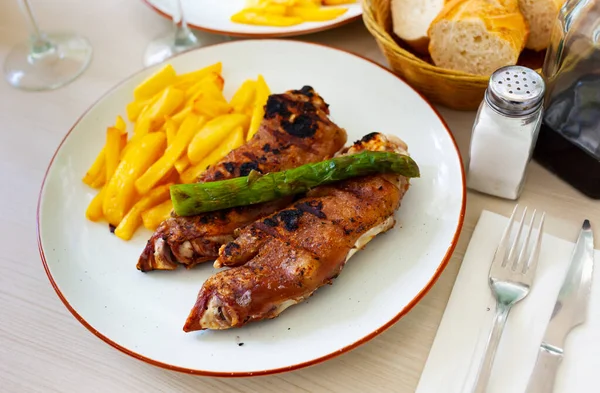 Close up of grilled pigs trotter with fried potato and asparagus served on white plate on wooden table — Stock Photo, Image