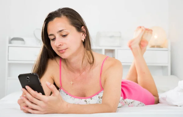 Smiling girl resting and using smartphone on bed in bedroom — Stock Photo, Image