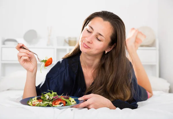 Женщина в синем шелковом халате ест салат в постели — стоковое фото