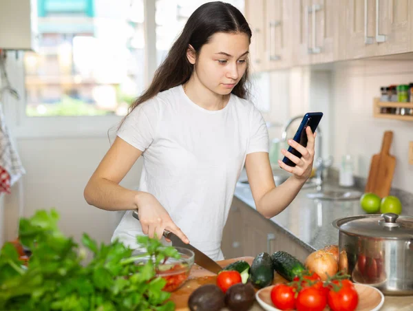 Positiv hemmafru matlagning grönsakssoppa och använda smartphone i köket — Stockfoto