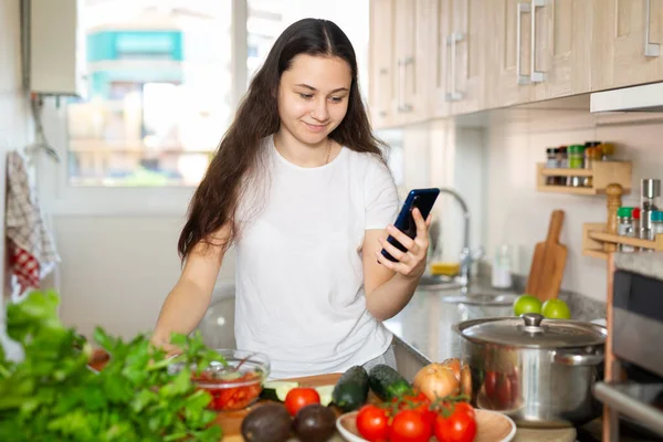Positiv hemmafru matlagning grönsakssoppa och använda smartphone i köket — Stockfoto