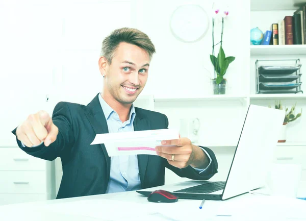 男人坐在笔记本电脑前，手里拿着纸质文件 — 图库照片