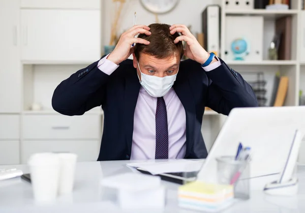 Annoyed worker wearing medical facial mask engaged in business activities at table in office