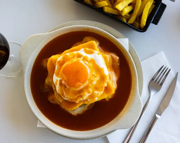 Hearty Portuguese lunch Francesinha, sandwich with bread, ham, fresh sausage, roasted meat, melted cheese, with fried egg on top and warm beer and tomato sauce