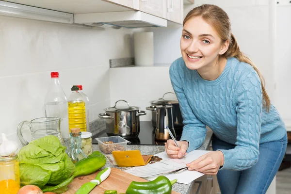 Young housewife filling banking documents and smiling in kitchen