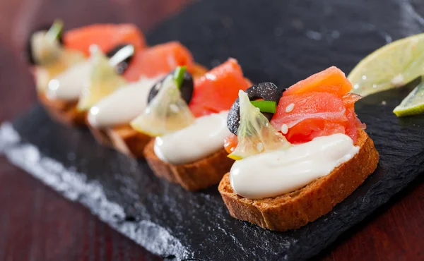 Canapes on toasted bread with smoked salmon, olives and creamy sauce on black serving board..