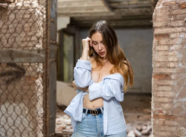 Portrait of young desirable woman taking off her shirt, demonstrating naked shoulders, covering her breast in abandoned building.