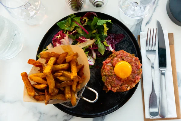 Traditional appetizer of French cuisine is beef tartare made from raw meat, capers, onions and egg yolk. Served with French ..fries and fresh herbs