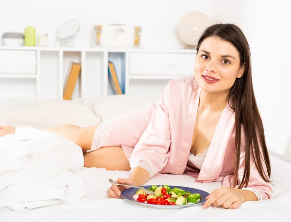 正かなり女性でバスローブを保持フォークと食べる野菜サラダでベッドに家 — ストック写真