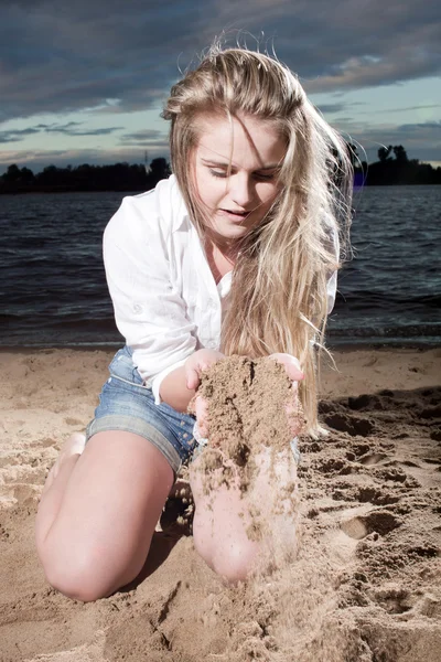 De jonge blonde zit op de kust en strews zand — Stockfoto