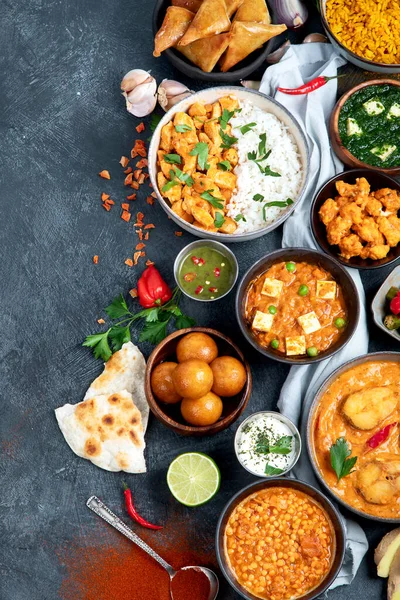 Dishes of indian cuisine. Curry, butter chicken, rice, lentils, paneer, samosa, spices. Bowls and plates with indian foodon dark on dark background. Top view with copy space.