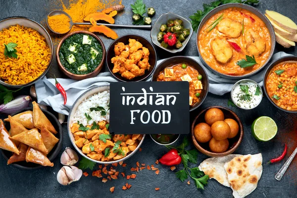 Dishes of indian cuisine. Curry, butter chicken, rice, lentils, paneer, samosa, spices. Bowls and plates with indian foodon dark on dark background. Top view.