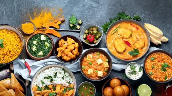 Dishes of indian cuisine. Curry, butter chicken, rice, lentils, paneer, samosa, spices. Bowls and plates with indian foodon dark on dark background.  Top view with copy space.