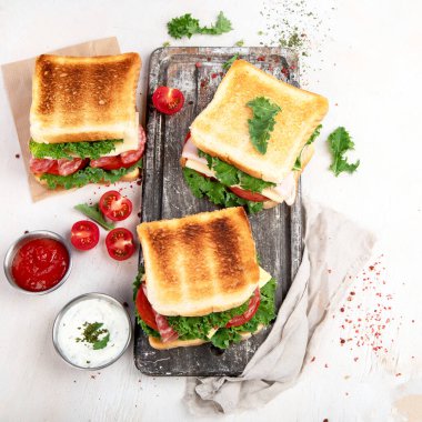 Sandwich with cheese, ham, tomato and fresh salad, made of whole wheat bread with a crispy crust on a wooden board with sauces on a white background, top view.