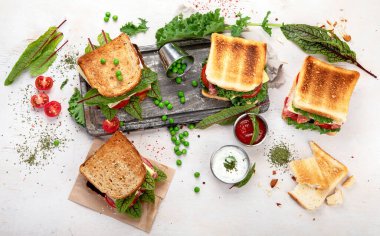Sandwich with cheese, ham, tomato and fresh salad, made of whole wheat bread with a crispy crust on a wooden board with sauces on a white background, top view.