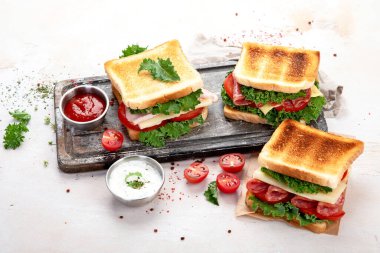 Sandwich with cheese, ham, tomato and fresh salad, made of whole wheat bread with a crispy crust on a wooden board with sauces on a white background, top view.