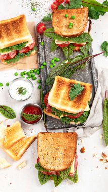 Sandwich with cheese, ham, tomato and fresh salad, made of whole wheat bread with a crispy crust on a wooden board with sauces on a white background, top view.
