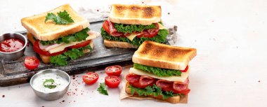 Sandwich with cheese, ham, tomato and fresh salad, made of whole wheat bread with a crispy crust on a wooden board with sauces on a white background, top view, panorama with copy space.