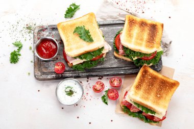 Sandwich with cheese, ham, tomato and fresh salad, made of whole wheat bread with a crispy crust on a wooden board with sauces on a white background, top view.