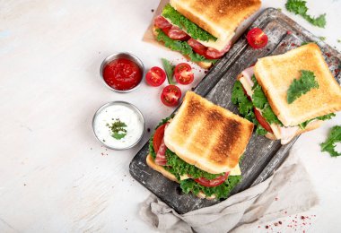 Sandwich with cheese, ham, tomato and fresh salad, made of whole wheat bread with a crispy crust on a wooden board with sauces on a white background, top view with copy space.