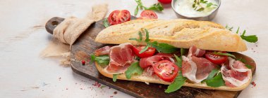 Baguette sandwich with prosciutto ham, cherry tomatoes and arugula on a board. White background, top view. Panorama.
