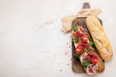 Baguette sandwich with prosciutto ham, cherry tomatoes and arugula on a board. White background, top view with copy space.