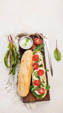 Preparing a fresh crusty baguette with cherry tomatoes, arugula and mozzarella cheese on a wooden board with sauce. White background. Top view.