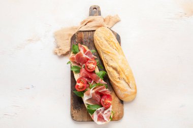 Baguette sandwich with prosciutto ham, cherry tomatoes and arugula on a board. White background, top view.