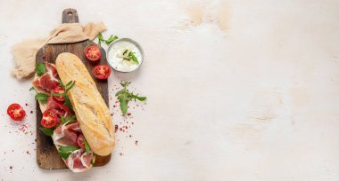 Baguette sandwich with prosciutto ham, cherry tomatoes and arugula on a board. White background, top view. Panorama with copy space.