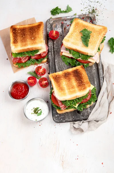 Sandwich with cheese, ham, tomato and fresh salad, made of whole wheat bread with a crispy crust on a wooden board with sauces on a white background, top view.