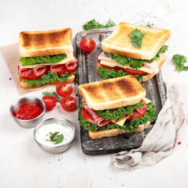 Sandwich with cheese, ham, tomato and fresh salad, made of whole wheat bread with a crispy crust on a wooden board with sauces on a white background, top view.