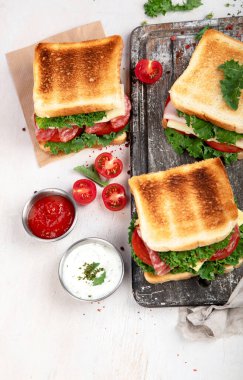 Sandwich with cheese, ham, tomato and fresh salad, made of whole wheat bread with a crispy crust on a wooden board with sauces on a white background, top view.