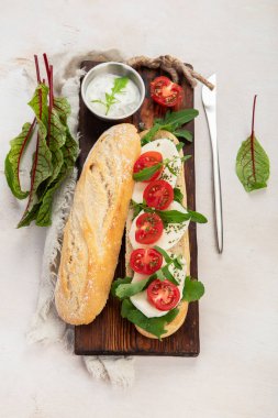 Preparing a fresh crusty baguette with cherry tomatoes, arugula and mozzarella cheese on a wooden board with sauce. White background. Top view.