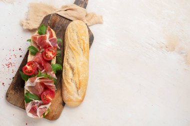 Baguette sandwich with prosciutto ham, cherry tomatoes and arugula on a board. White background, top view with copy space.