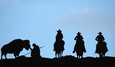 Silhouettes of three cowboys on horseback and an Indian with a buffalo against the blue sky on the arch of the entrance to Pullman City