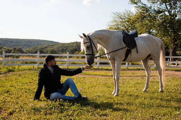 Mann sitzt auf dem Gras und füttert ein Pferd — Stockfoto