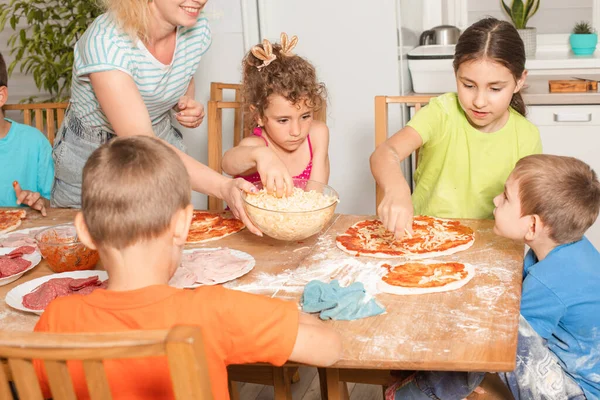 Boldog gyerekek ülnek egy asztalnál a konyhában, és pizzát csinálnak. — Stock Fotó