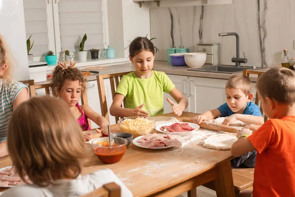 Niños felices se sientan en una mesa en la cocina y hacen pizza —  Fotos de Stock