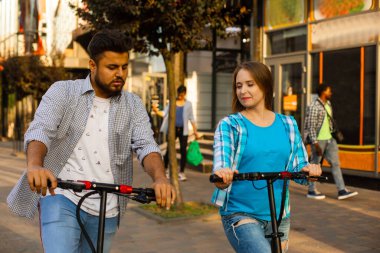 Sevgili çift elektrikli scooterlara biniyor ve birbirlerine bakıyorlar.