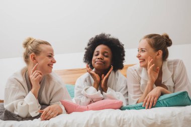 Genç kadınlar bornozlarıyla yatakta uzanıyor ve iletişim kuruyorlar. Mutlu kızlar spa otelinde dinleniyor.